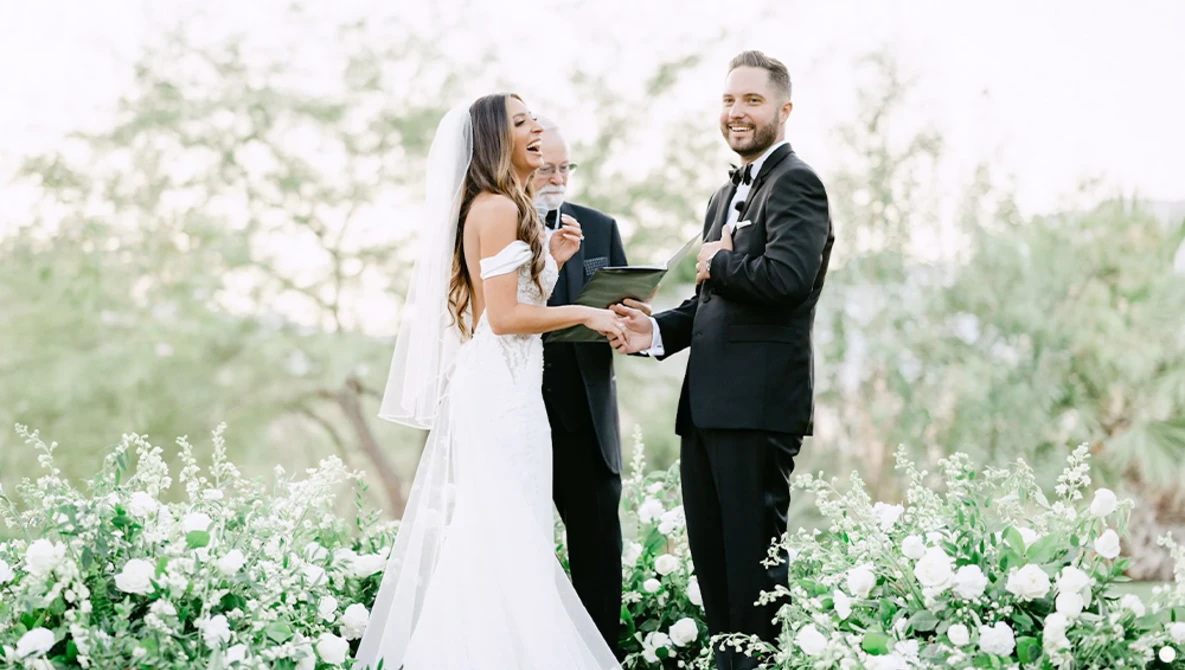 Bride and groom exchange vows during outdoor ceremony surrounded by white flowering plants.