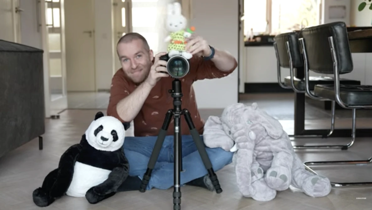 Photographer holding a telephoto lens on a tripod while posing with stuffed animals in an indoor studio setting.