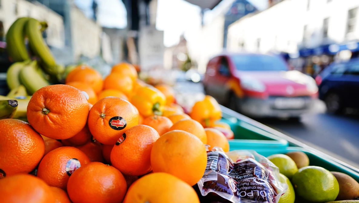 Fresh oranges and bananas displayed at an outdoor market stall with blurred urban street and parked cars in the background.