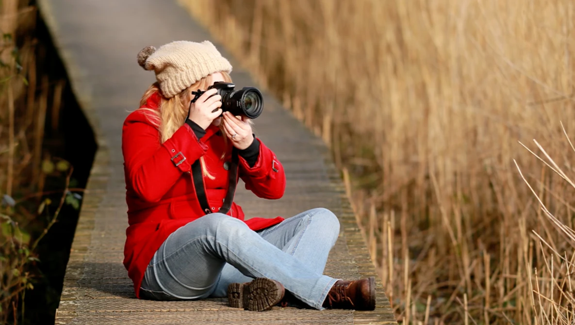 Photographer sitting on a wooden boardwalk holding a camera to their face, wearing a red jacket and cream beanie.