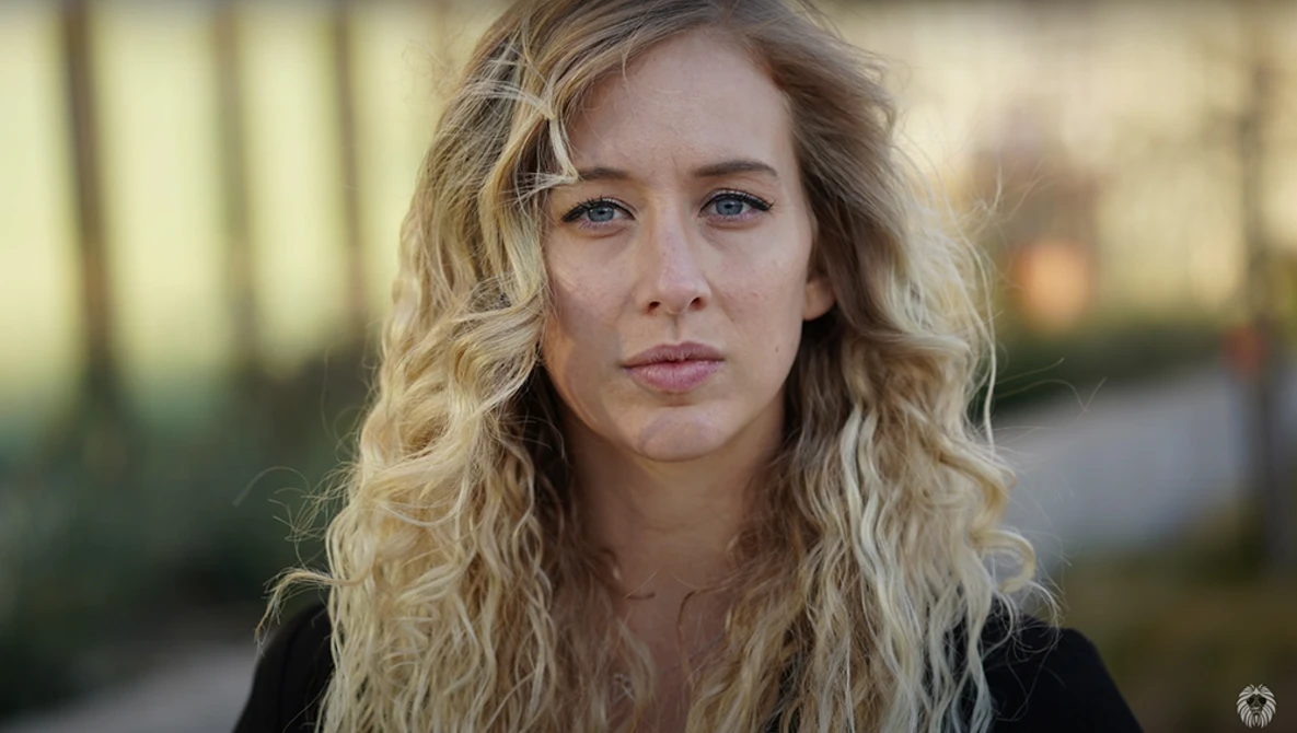 Woman with wavy blonde hair looking directly at camera with neutral expression outdoors.