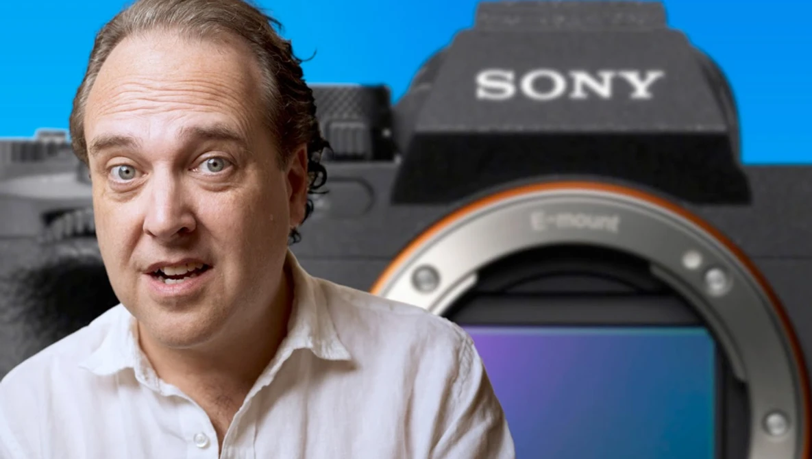 Man in white shirt gesturing expressively toward Sony mirrorless camera against blue background.