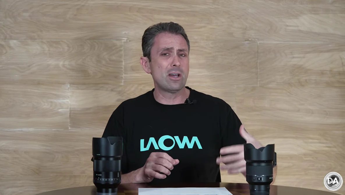 Man in black shirt with turquoise text speaking at table with two camera lenses positioned on either side.