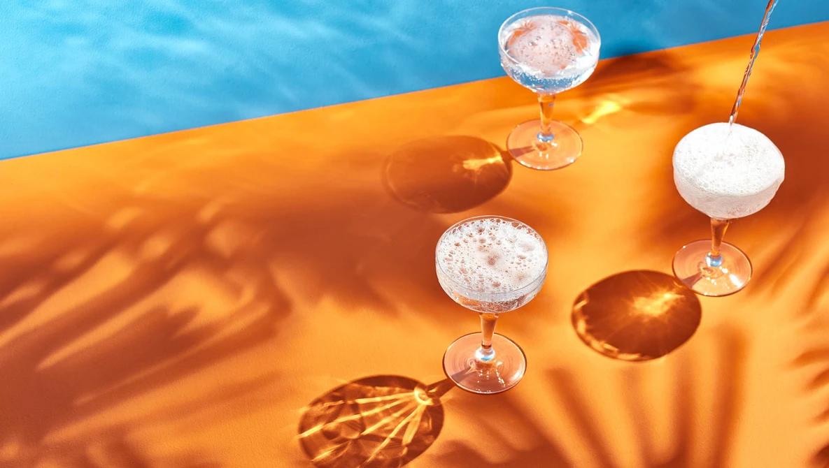 Cocktails suspended above an orange fabric with blue water and palm leaf shadows in bright sunlight.