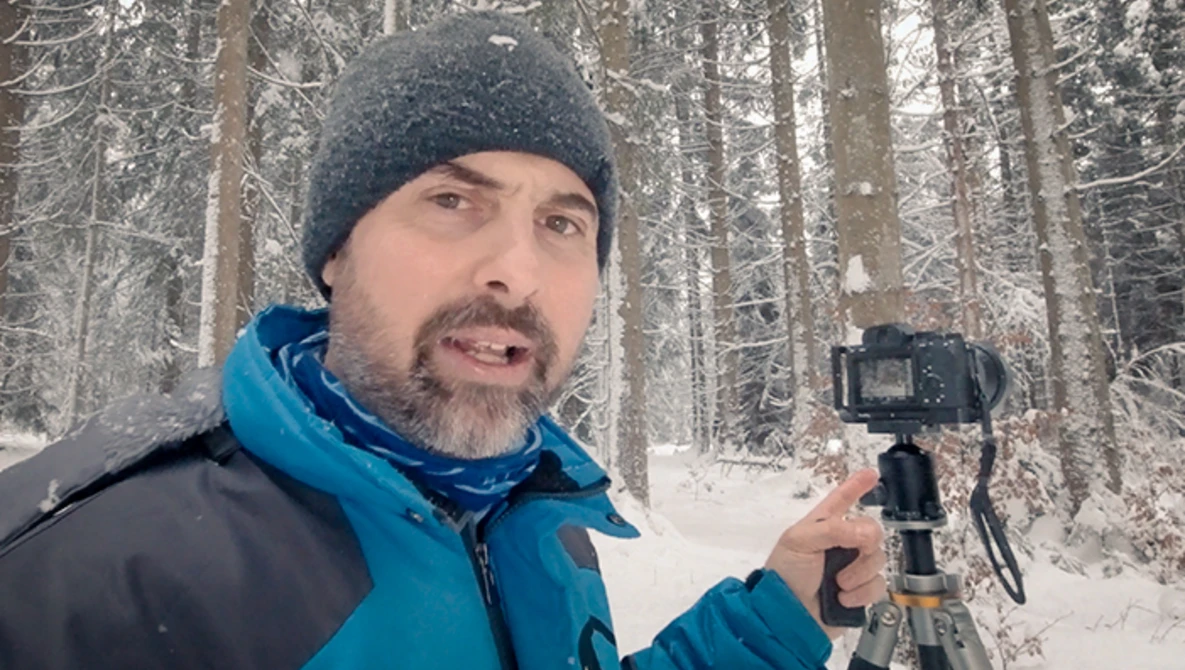 Videographer holding a gimbal stabilizer while filming in a snowy forest setting.