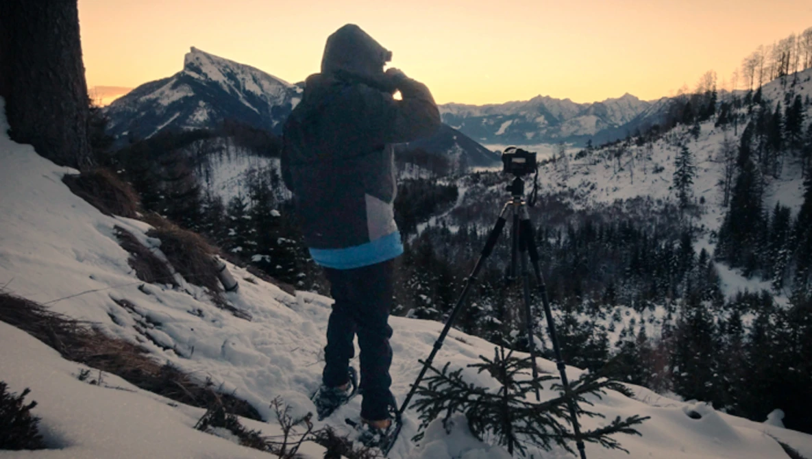 Photographer with camera on tripod overlooking snow-covered mountain landscape at golden hour.