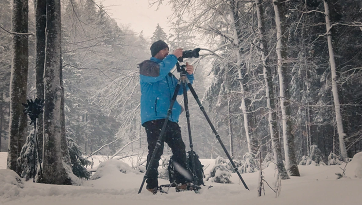 Photographer in blue jacket shooting with telephoto lens on tripod in snowy forest.