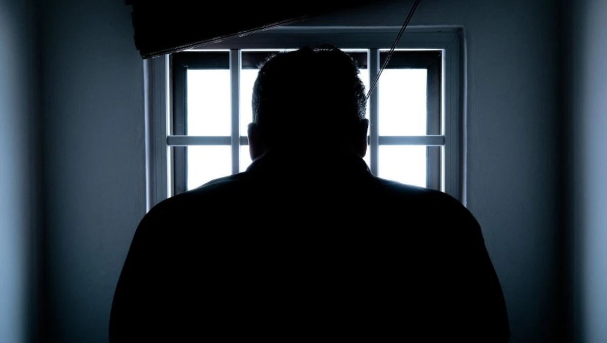 Silhouetted figure facing a bright window with gridded panes in a dimly lit interior space.
