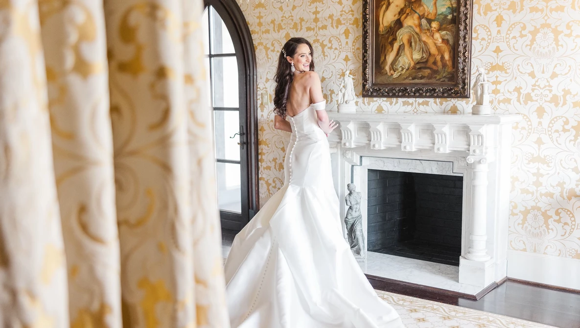 Bride in off-shoulder wedding dress with train standing by ornate fireplace in classical interior.