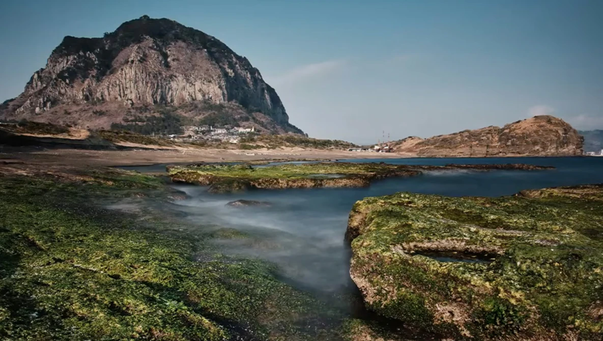 Moss-covered rocky shoreline with calm blue water and a dramatic peaked mountain rising behind a coastal settlement.