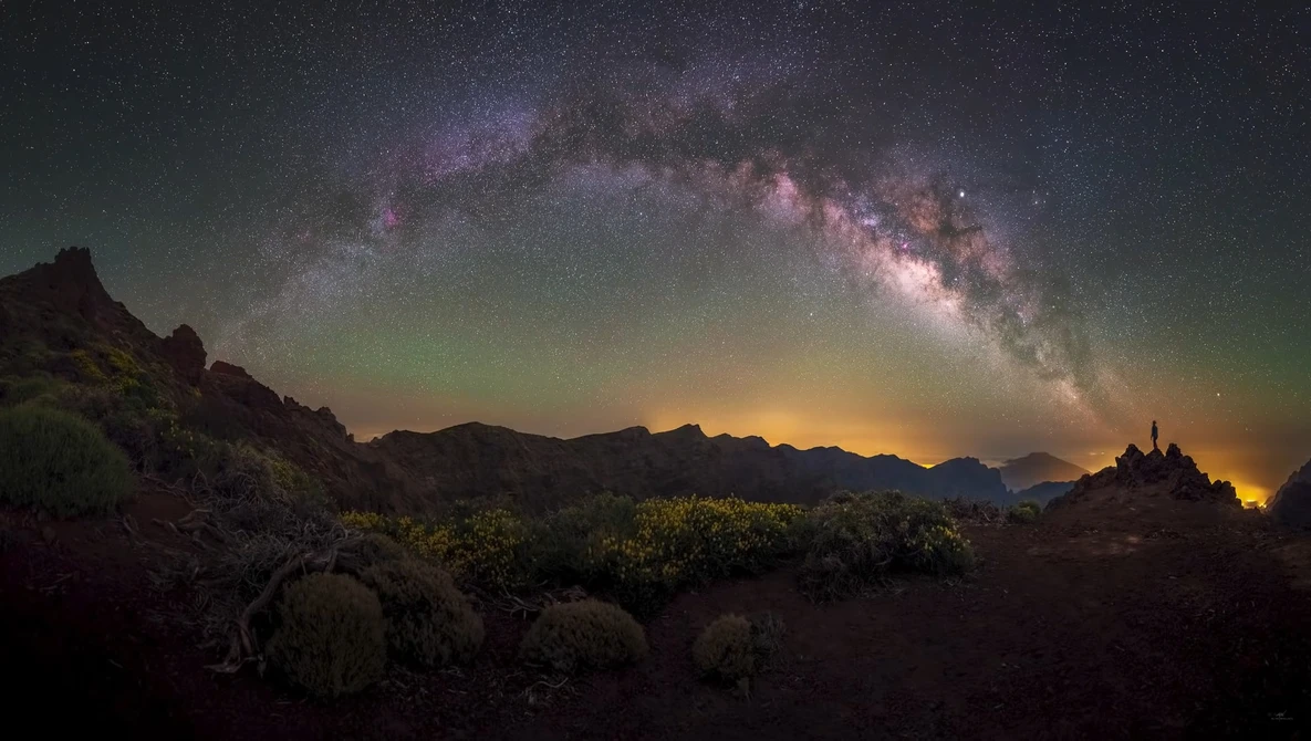 Milky Way galaxy arcing across night sky above desert landscape with mountains and distant lights.