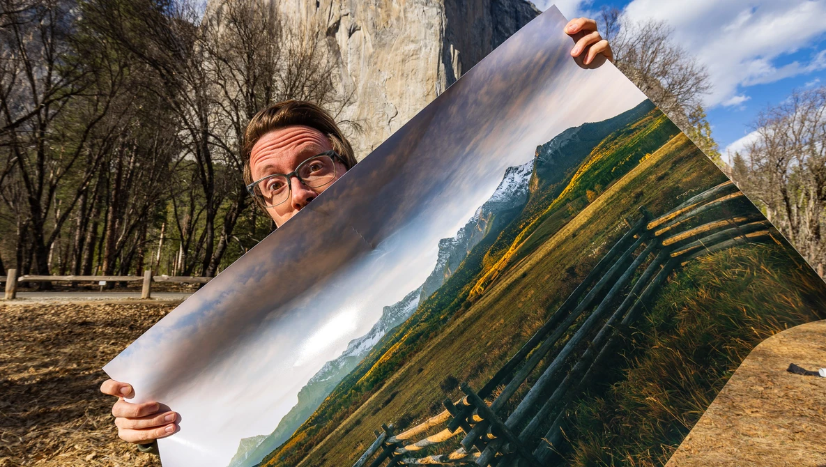 Photographer holding a printed landscape photograph outdoors, comparing the print to the actual mountain valley scene behind it.