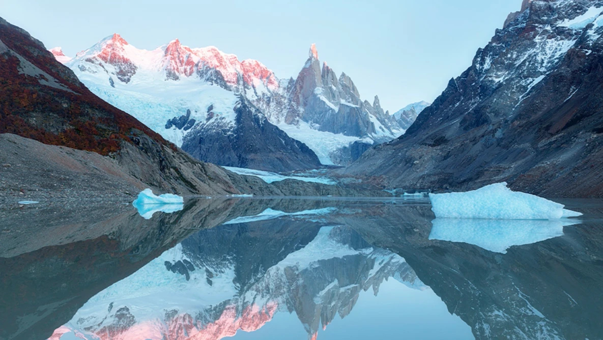 Alpine glacier lake with perfect mirror reflection of snow-capped peaks and icebergs at dawn.