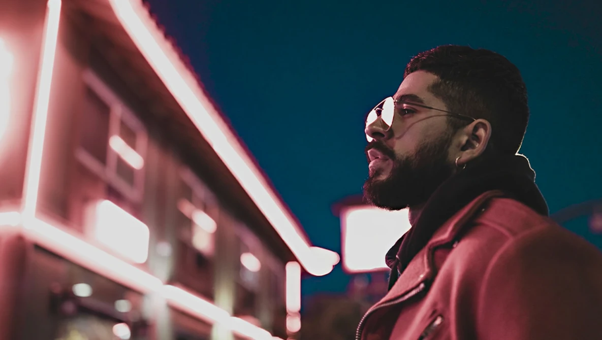 Man wearing glasses and maroon jacket against neon pink signage and teal night sky.