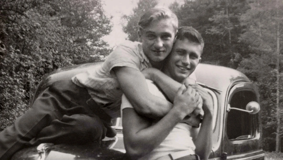 Two men posing playfully on the hood of a vintage automobile in a wooded setting.
