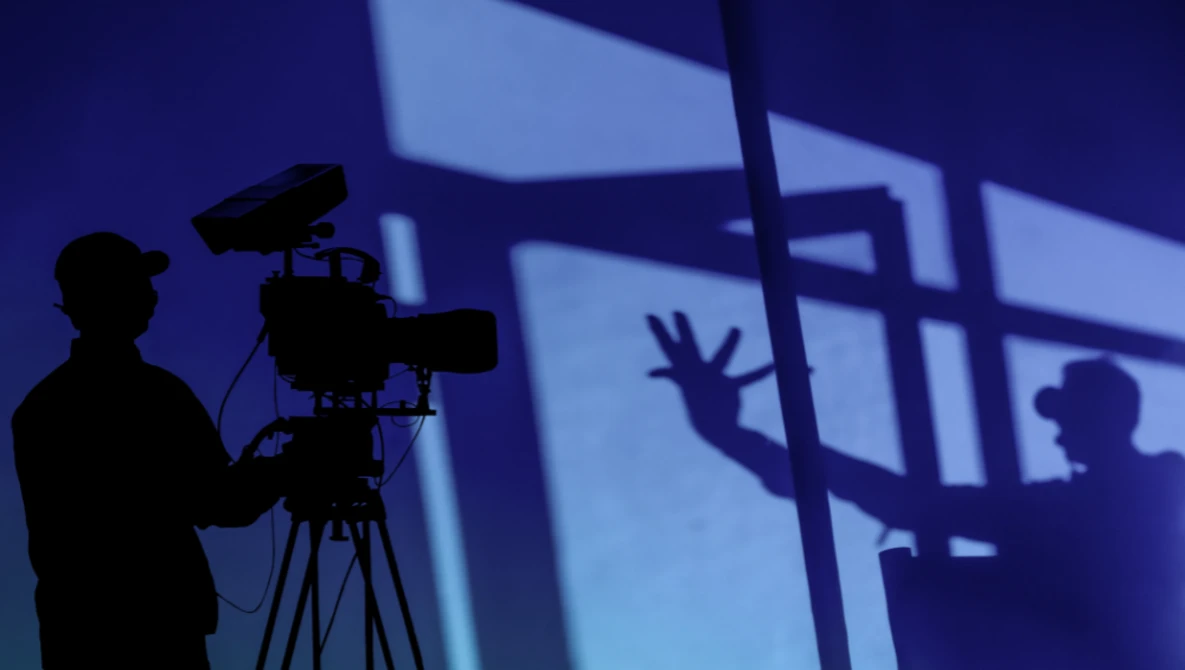 Silhouette of a filmmaker operating a professional video camera on a tripod, with shadows cast on a blue-lit wall behind.