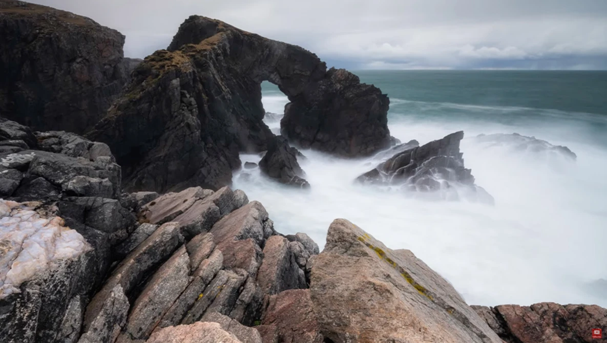 Rocky coastal outcrop with natural stone arch framing turbulent ocean swells and misty waves.