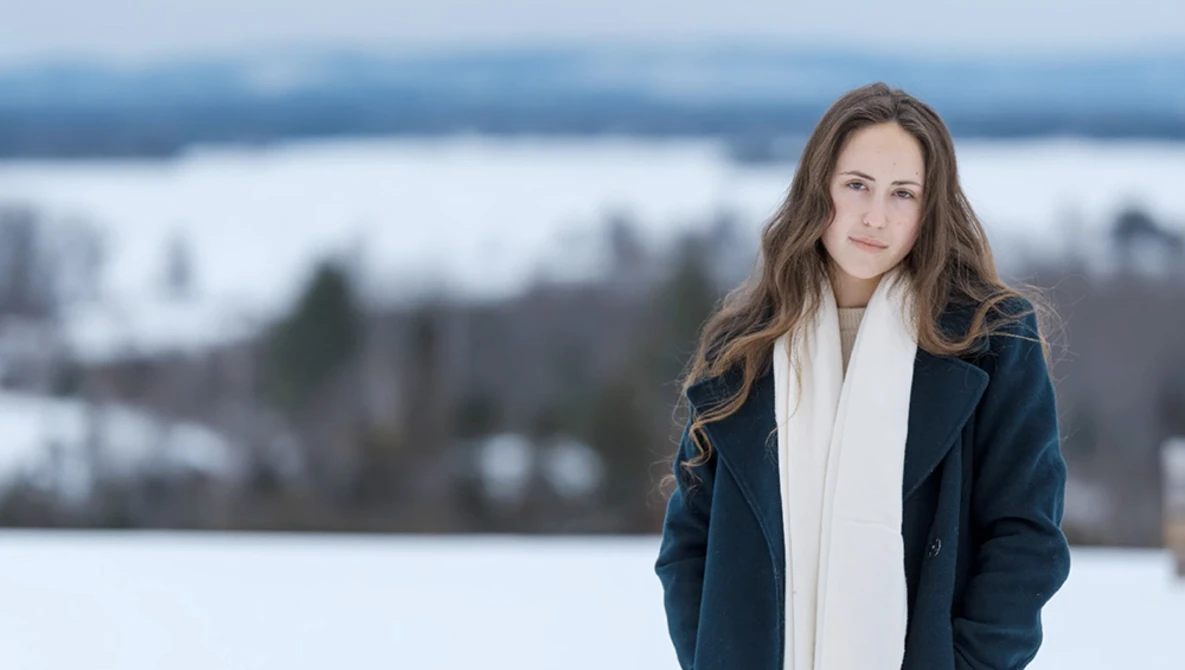Woman in dark jacket and white scarf standing outdoors with snowy landscape behind her.