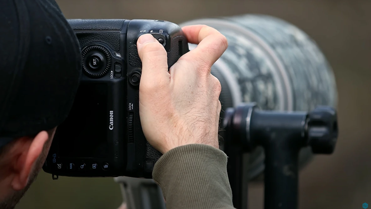 Photographer holding a Canon mirrorless camera in shooting position, with telephoto lens visible in background.