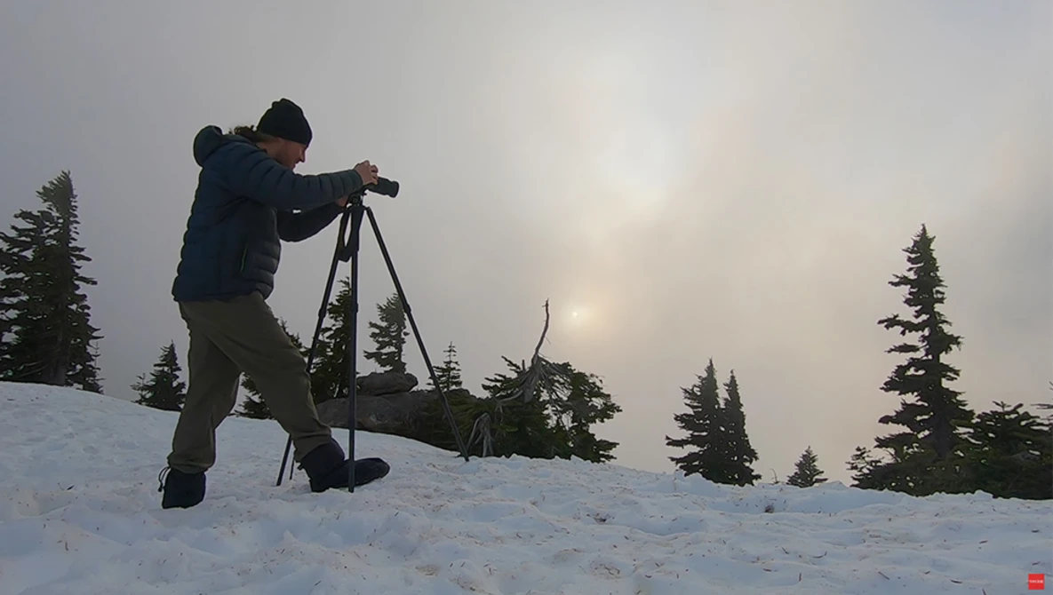 Photographer using a tripod-mounted telescope on snow-covered ground with coniferous trees and overcast sky.