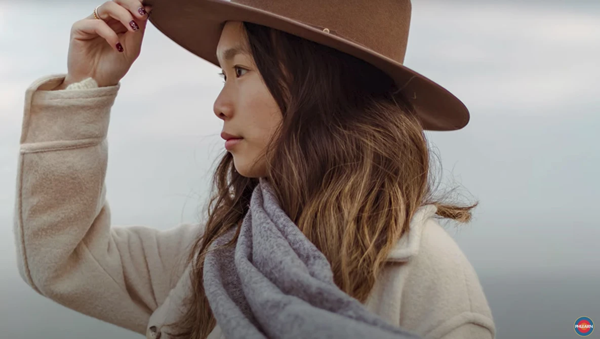 Woman in cream sweater and brown hat adjusting her hat brim while looking to the side.