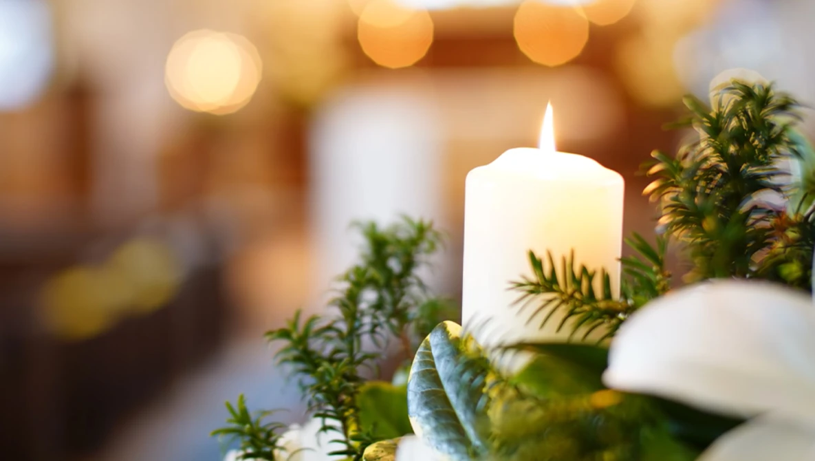 Lit white pillar candle surrounded by green foliage and white flowers with warm bokeh lights in soft focus background.