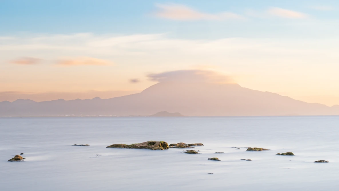 Volcanic mountain at sunrise across calm water with scattered rocks in foreground.