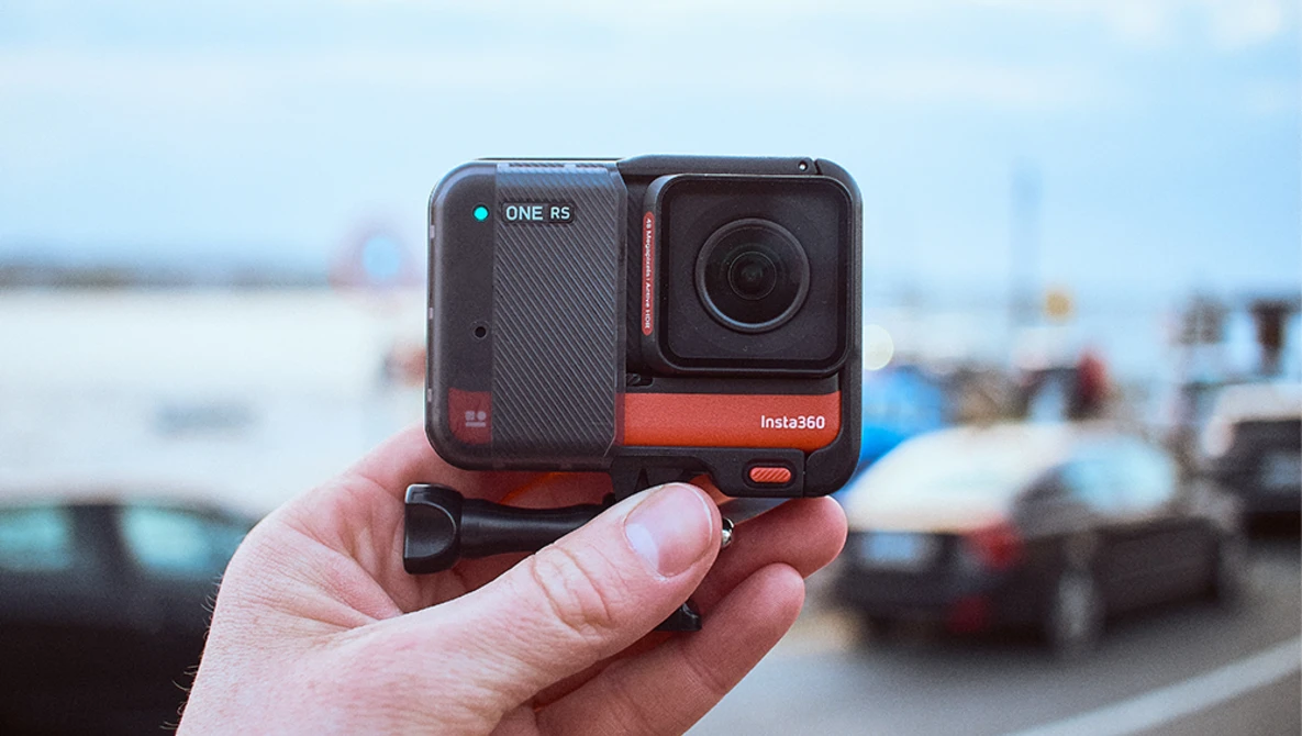 Hand holding an Insta360 ONE RS action camera with dual-lens setup against a blurred outdoor parking lot background.