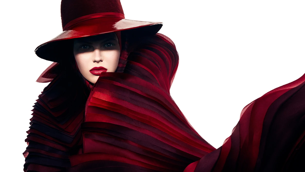Portrait of a person wearing a dramatic red and burgundy draped garment and wide-brimmed hat against white background.
