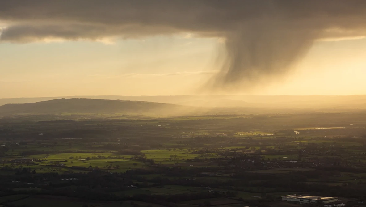 Aerial landscape with rolling hills, patchwork fields, and dramatic storm clouds illuminated by golden sunlight.