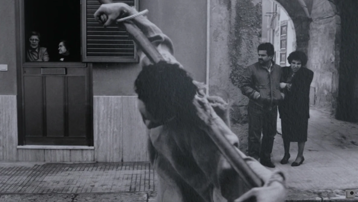 Black and white film still showing a figure suspended mid-air in an urban alleyway while pedestrians observe from nearby.