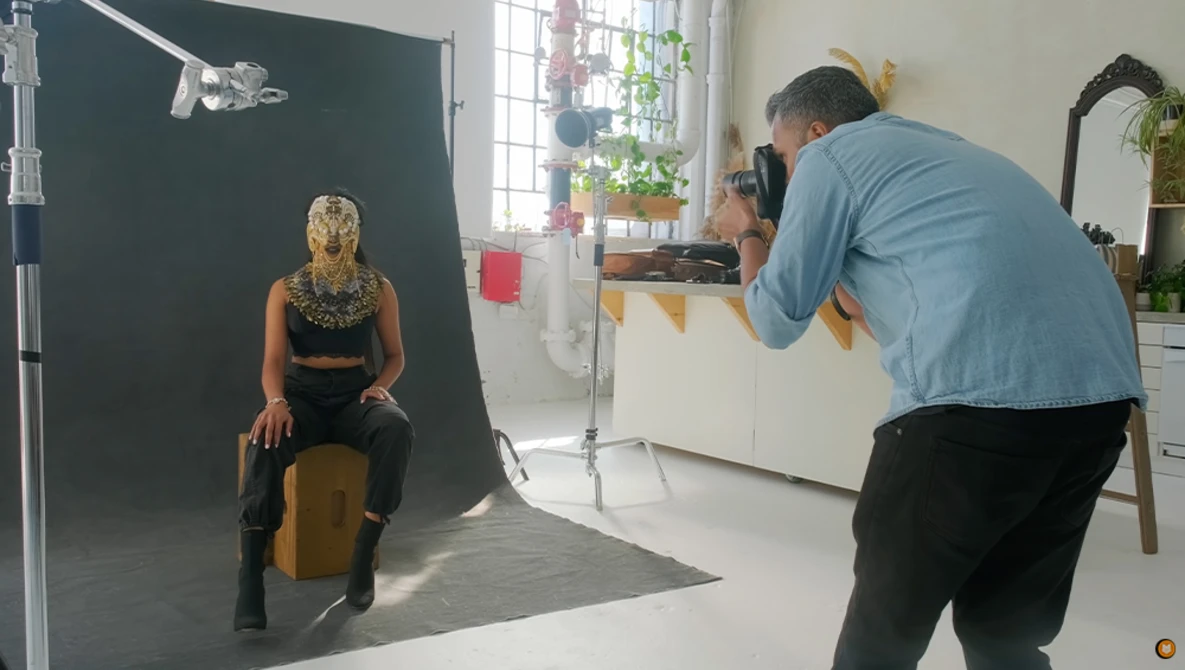 Photographer shooting portrait of woman seated on wooden stool against dark backdrop in studio.