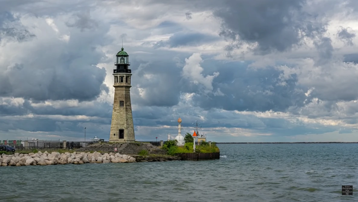 Stone lighthouse with green cupola on rocky breakwater extending into calm waters under dramatic cloudy sky.