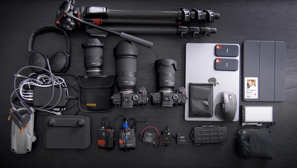 Flat lay of professional video and photography gear including cameras, lenses, audio equipment, and accessories arranged on dark surface.