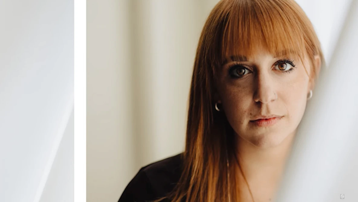 Woman with auburn hair and bangs looking directly at camera against neutral background.