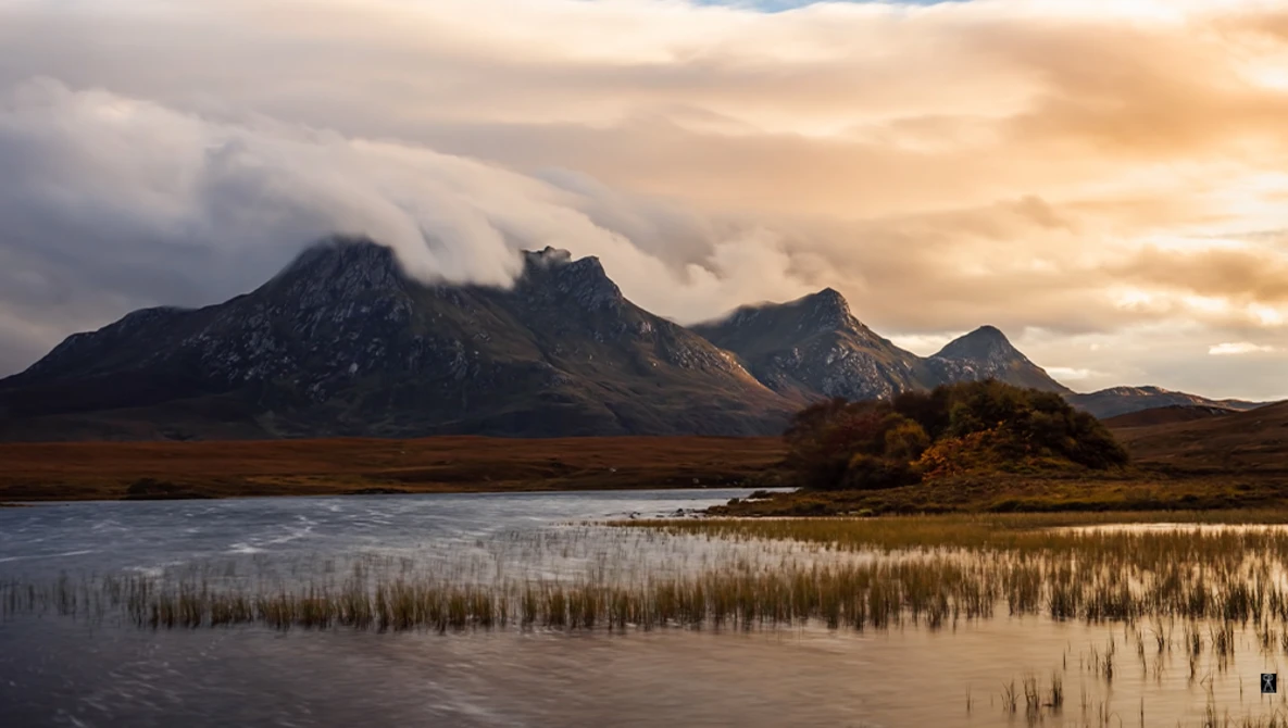 Mountain peaks with low-lying clouds reflected in a still wetland at golden hour.