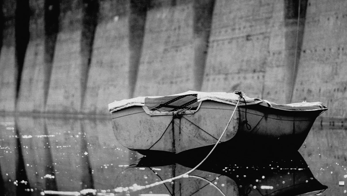 Small fishing boat with furled sail moored against a concrete dam wall, photographed in black and white.