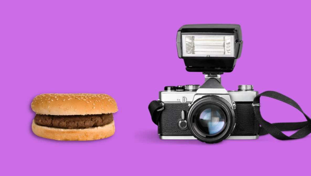 A hamburger and a vintage rangefinder camera with flash unit side by side on a magenta background.
