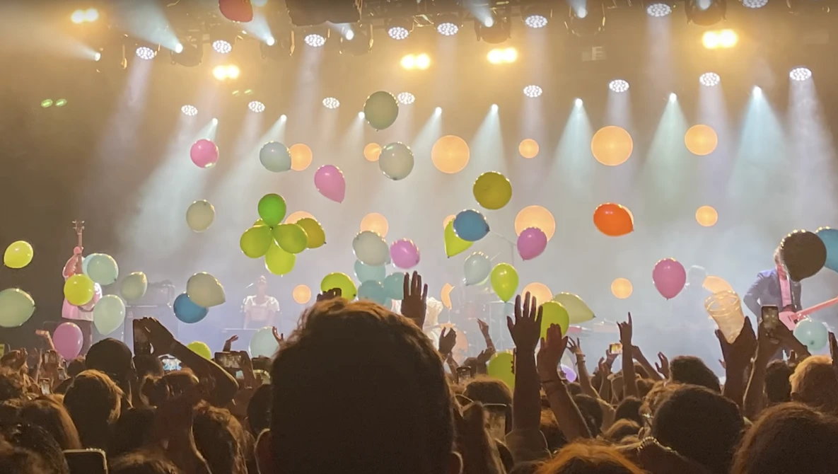 Silhouetted crowd with raised hands at concert with suspended colorful balloons and dramatic stage lighting.