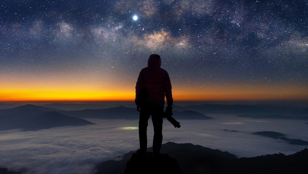 Silhouetted figure standing on a mountain peak beneath the Milky Way galaxy at twilight.