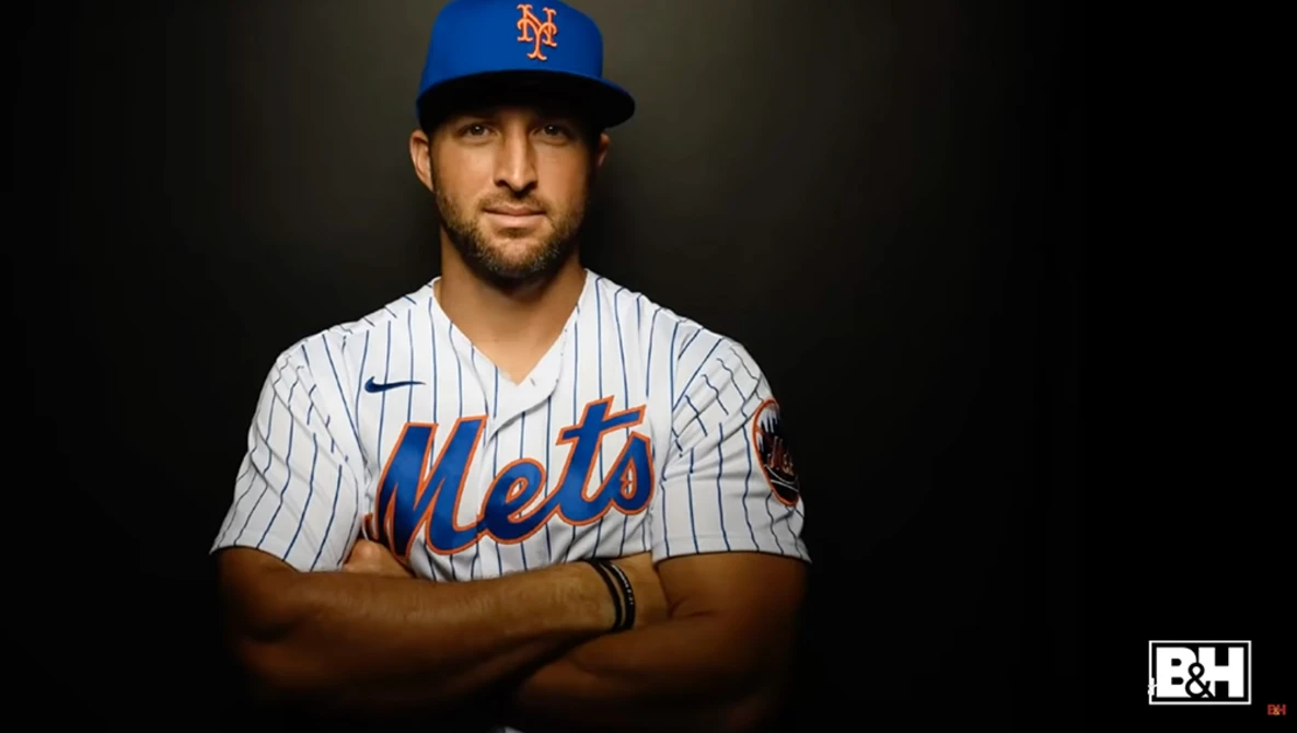 Baseball player in New York Mets pinstriped uniform and blue cap, arms crossed, against dark background.