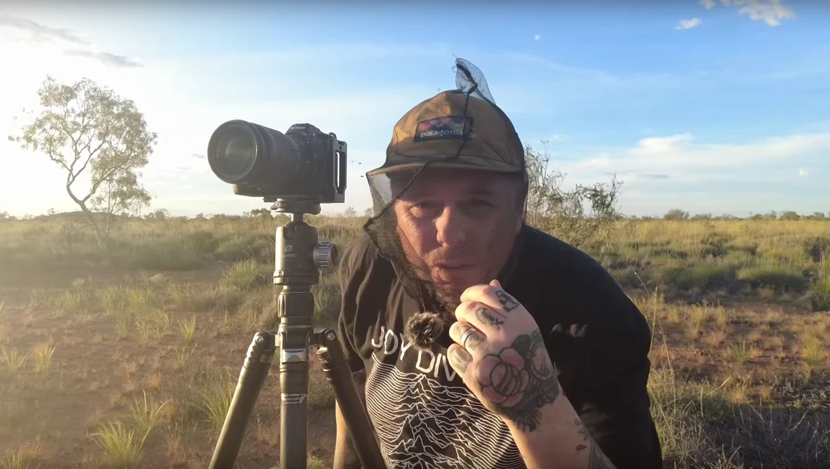 Photographer posing with telephoto lens mounted on tripod in open grassland at golden hour.