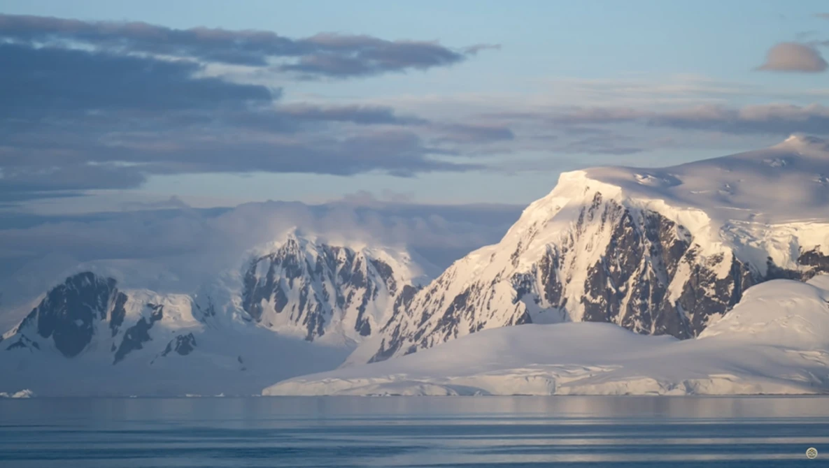 Snow-covered mountain peaks rising from calm blue water under a partly cloudy sky.