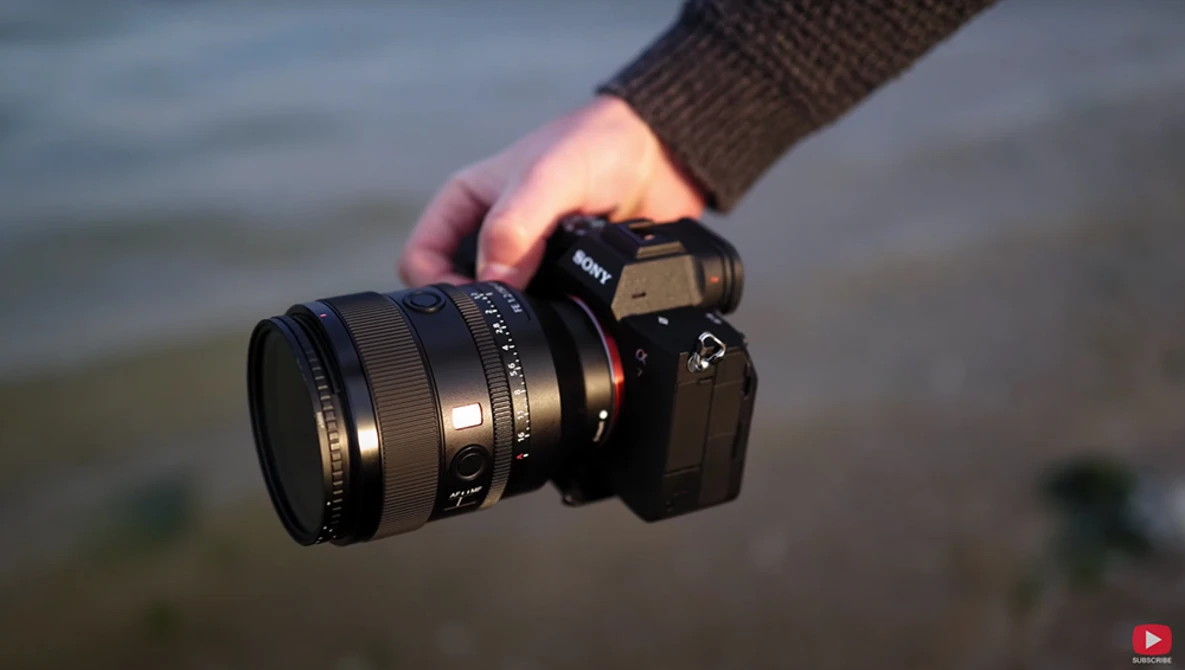 Photographer holding a mirrorless camera with prime lens, backlit by water and sky.