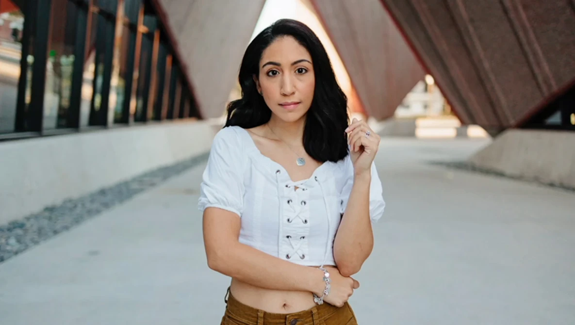 Woman in white lace-up shirt and tan pants holding white hood in architectural hallway.