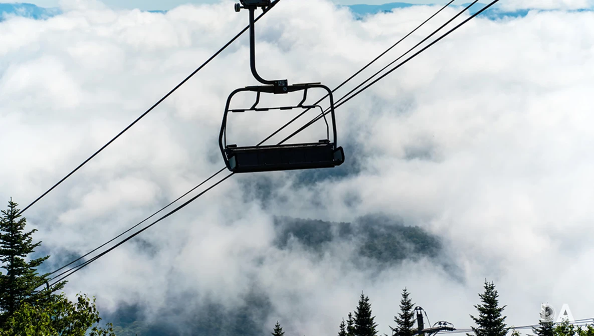 Ski lift chair suspended above clouds with forested mountains partially shrouded in mist below.