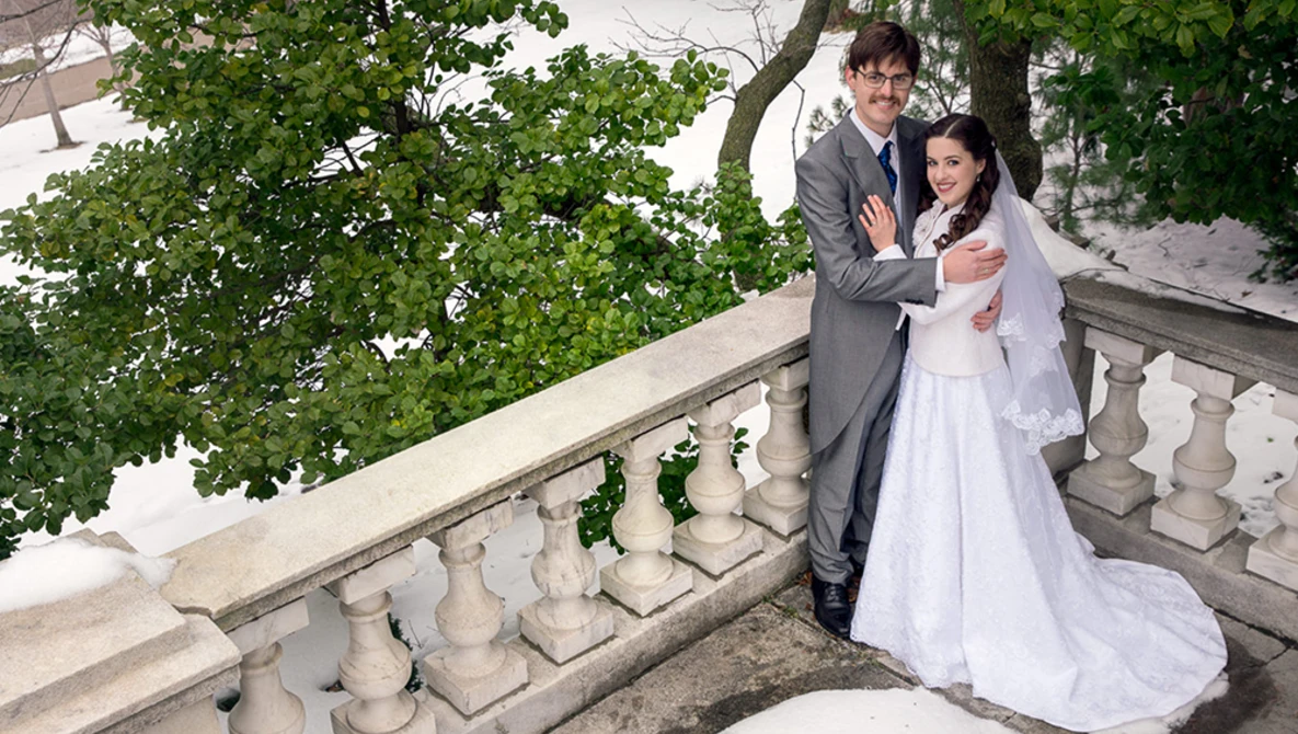 Bride and groom posing together on a stone balustrade with lush green foliage and trees in the background.