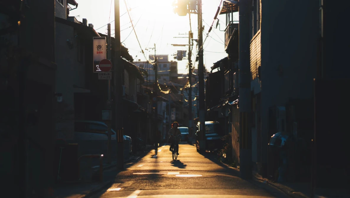 Cyclist riding alone down a narrow urban alley at golden hour with strong backlit sun creating dramatic shadows.