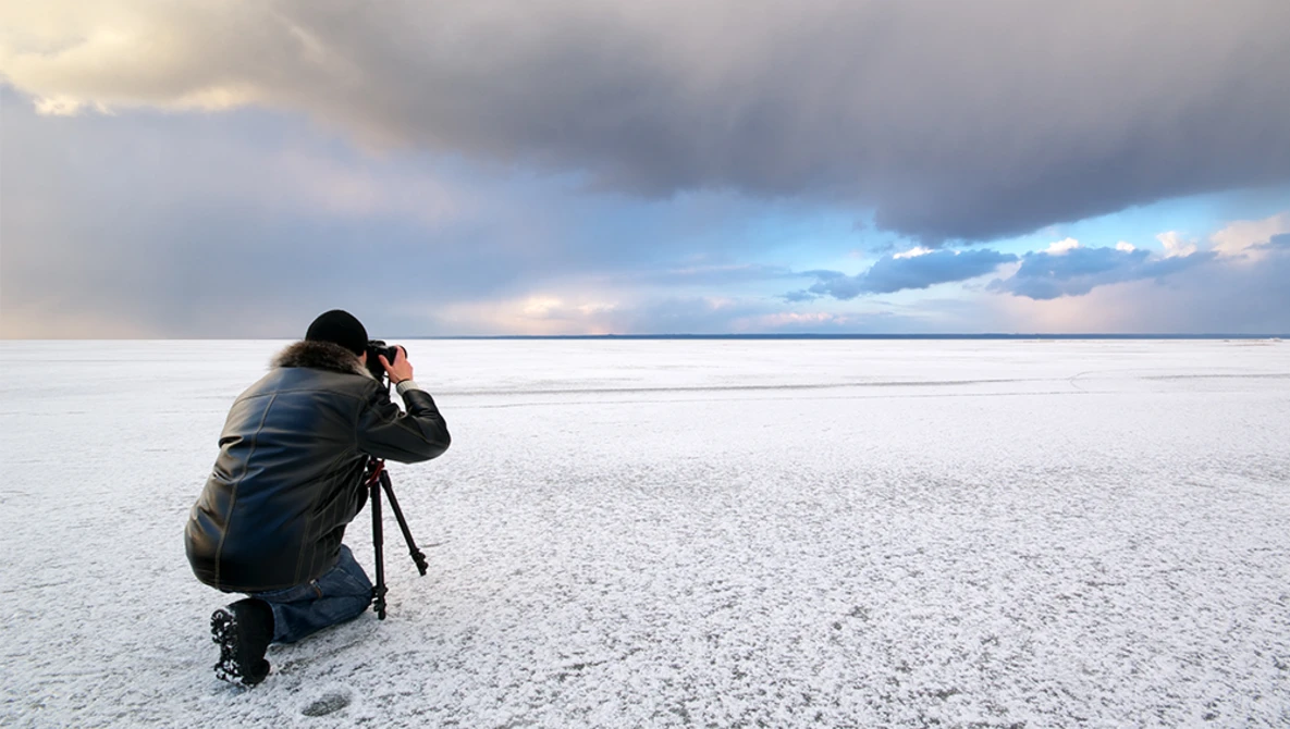 Photographer crouching on a vast salt flat, camera raised to eye level, capturing an expansive sky with dramatic cloud formations.