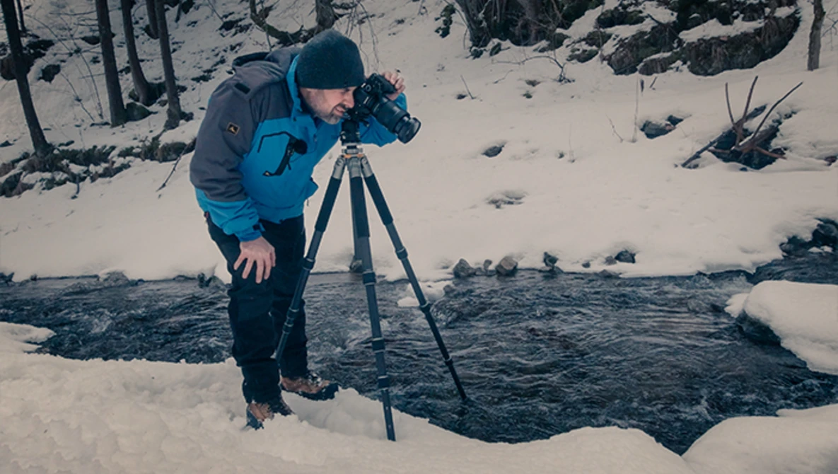Photographer operating a camera on tripod beside an unfrozen stream in a snow-covered forest.